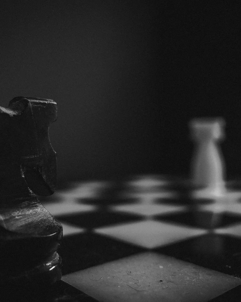 A black chess knight in sharp focus with a white rook out of focus in the background, on a dimly lit chessboard